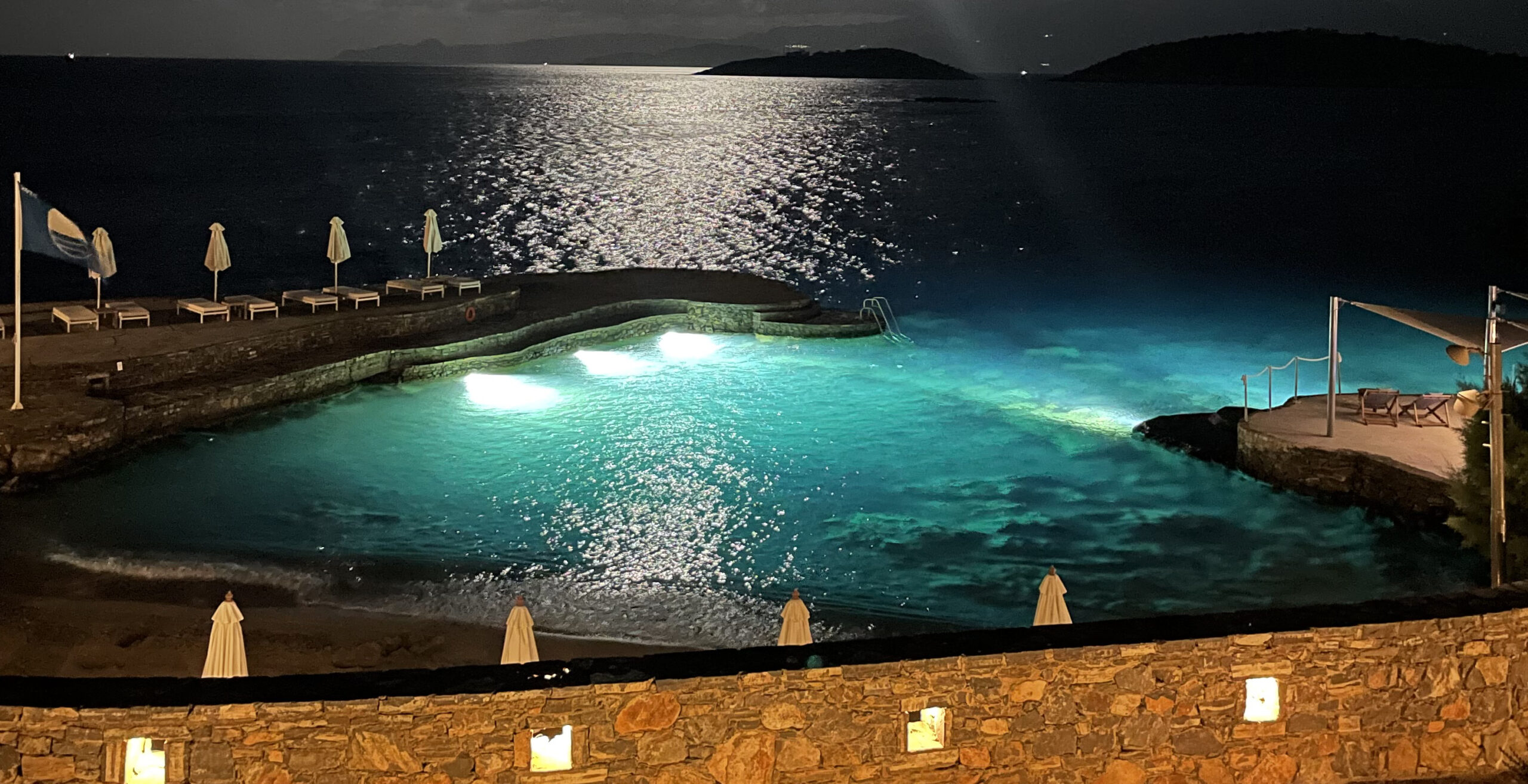 Ein Pool bei nach mit türkisblauem Wasser und einer Mosle mit Sonnenliegen und Sonnenschirmen.